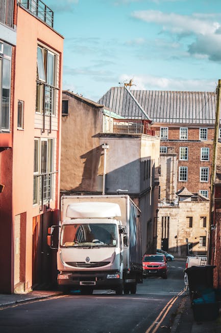 A white box truck parked on a narrow street in Islington, outside a multi-story residential building with a peach-colored facade and large windows, part of a home relocation or furniture transport pro