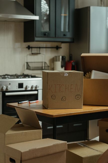 A young woman sitting on a wooden floor inside a room, surrounded by numerous large cardboard boxes of various sizes with black and red tape. She is wearing a white sleeveless top, beige pants, and bl
