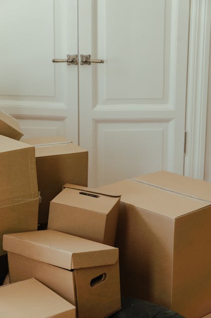 A person, partially visible, is seen arranging and stacking medium-sized cardboard boxes inside a room with light coming through a nearby window. The individual is wearing light-colored clothing and w