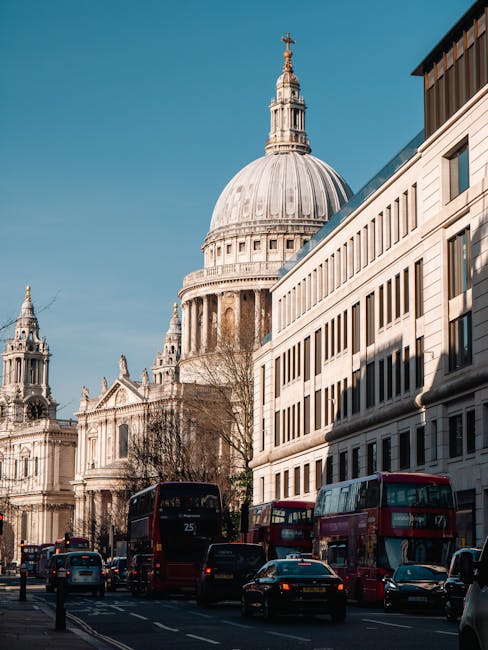 A wide city street scene in central London, featuring a mix of historic and modern buildings on both sides, with pedestrians walking along the pavement and vehicles, including red buses and cars, trav